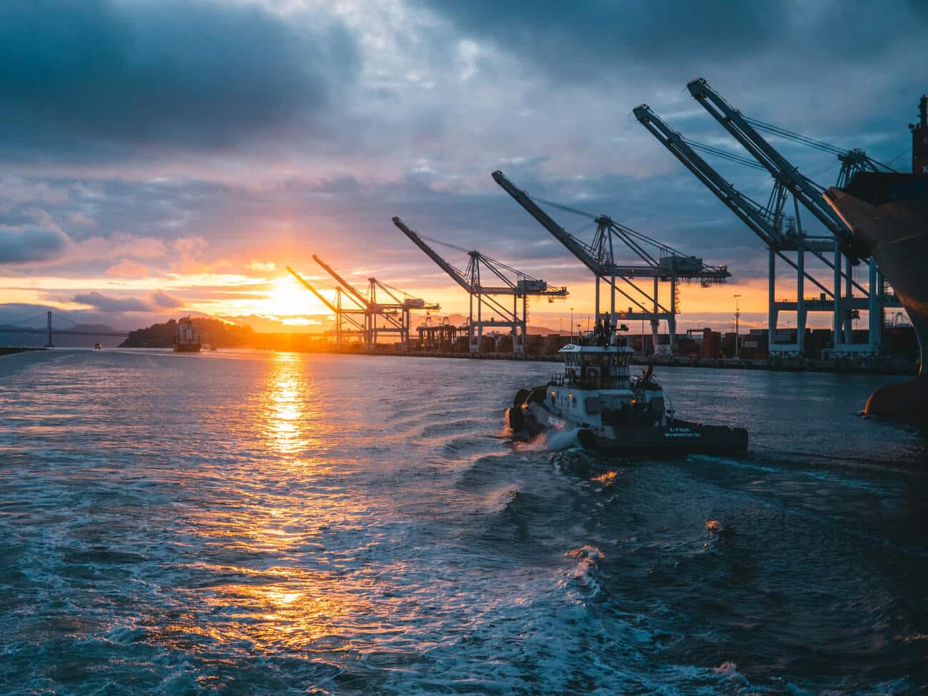 Operaciones marítimas de carga y descarga de hidrocarburos en puerto industrial al atardecer, con remolcador y grúas portuarias